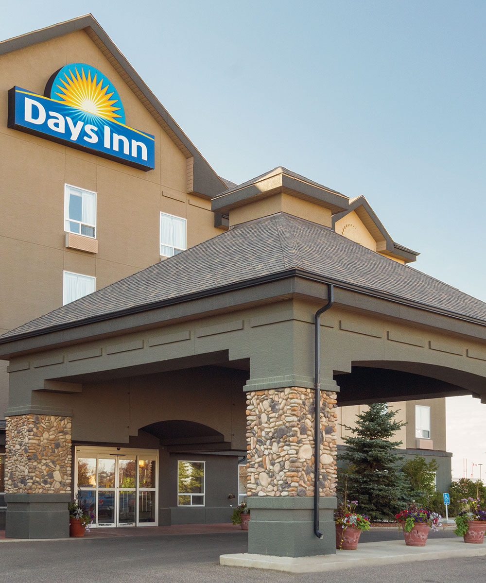 Entranceway to Days Inn Red Deer, Alberta featuring a large stone and concrete portico, green trees and larged pots of flowers placed throughout.
