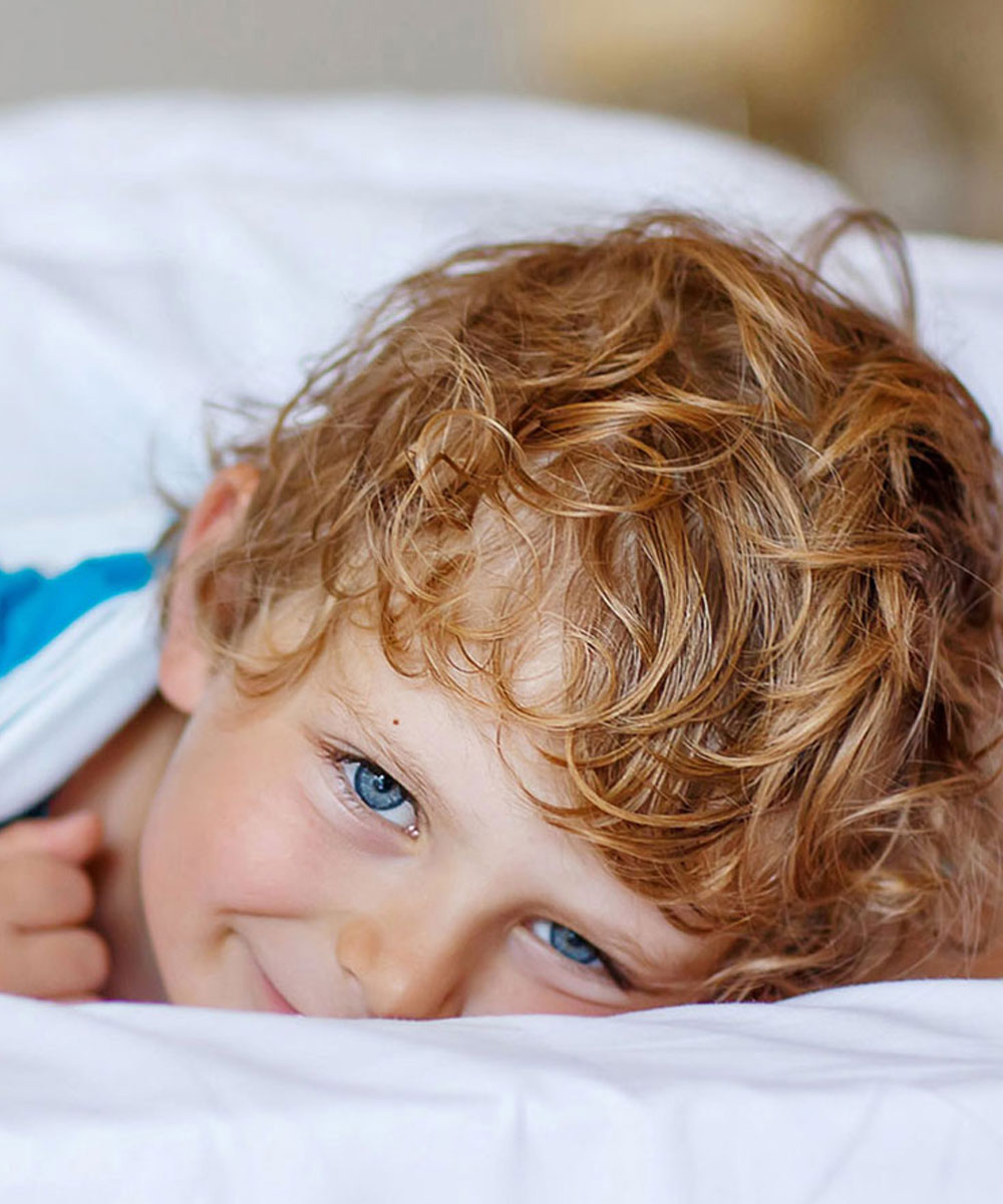 Photo of a smiling blonde blue eyed boy peering out from under white bed covers. 