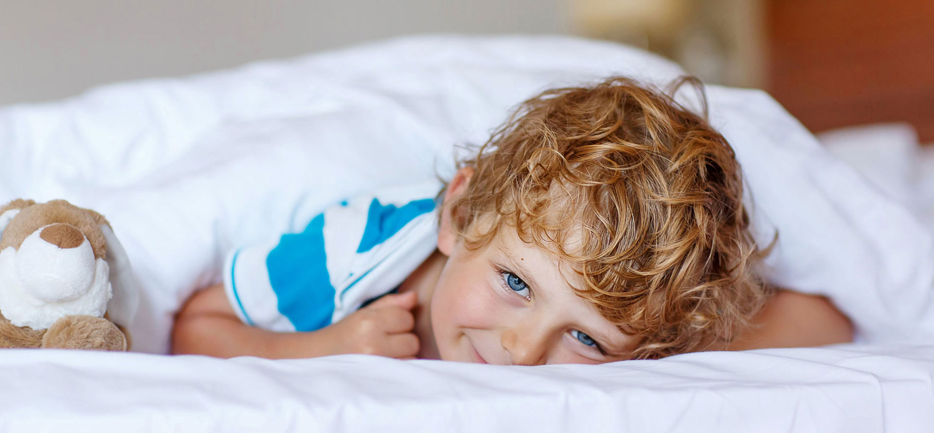 Large photo of a smiling blonde boy with bright blue eyes lying under white covers with a plush toy dog.