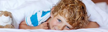 Small photo of a smiling boy with blonde hair and blue eyes peering out from under white bed covers.