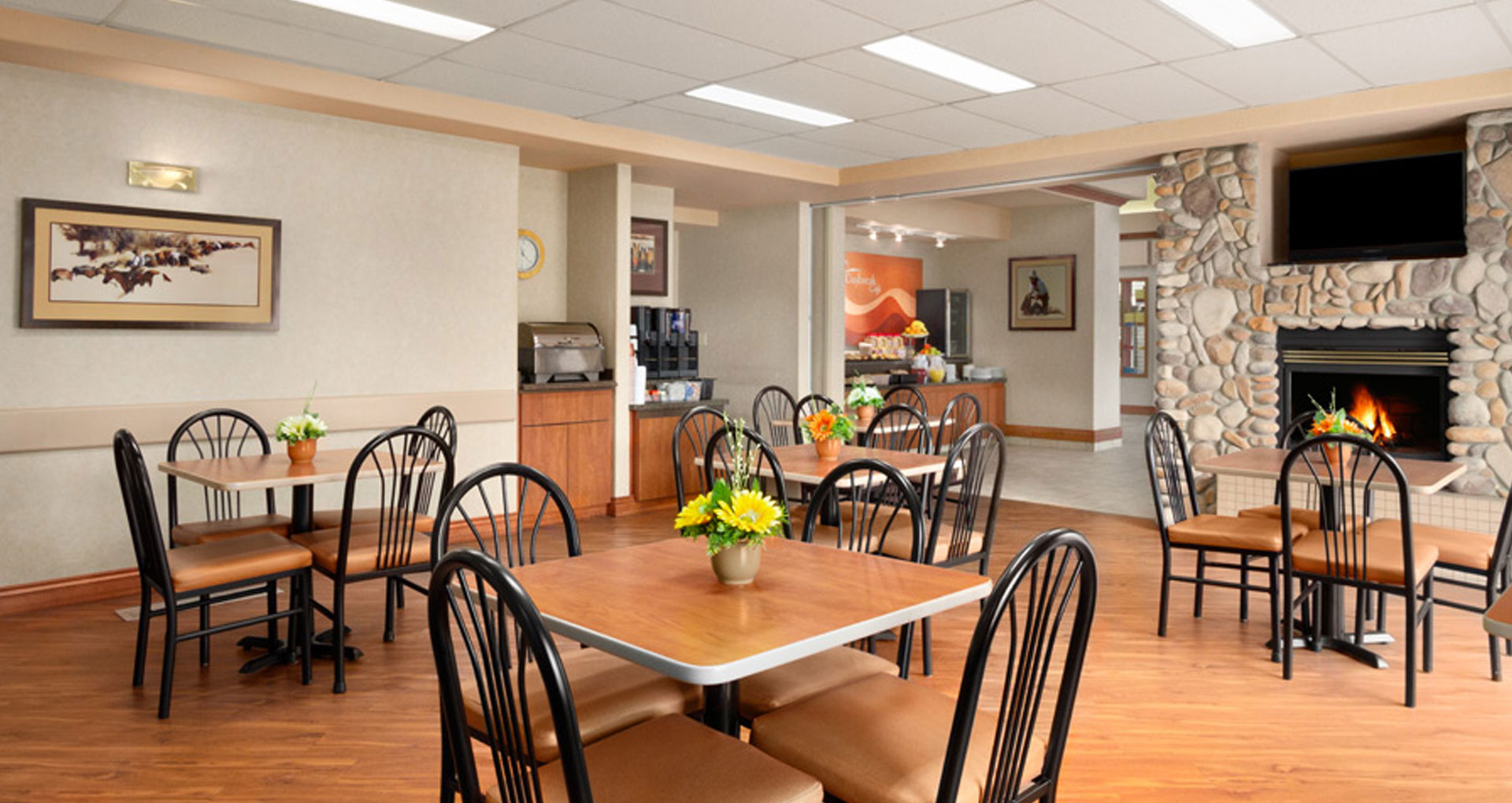 Large panoramic view of the Daybreak Café at Days Inn Red Deer with square eating tables and comfortable chairs are placed around a lit fireplace.
.
